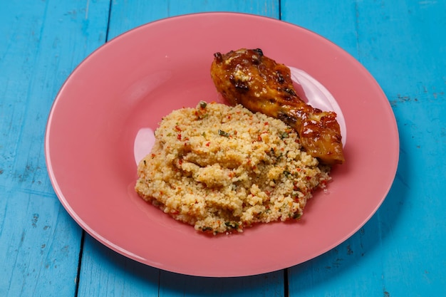 Foto cuscuz em molho e especiarias com coxa de frango grelhada em um prato sobre uma mesa azul