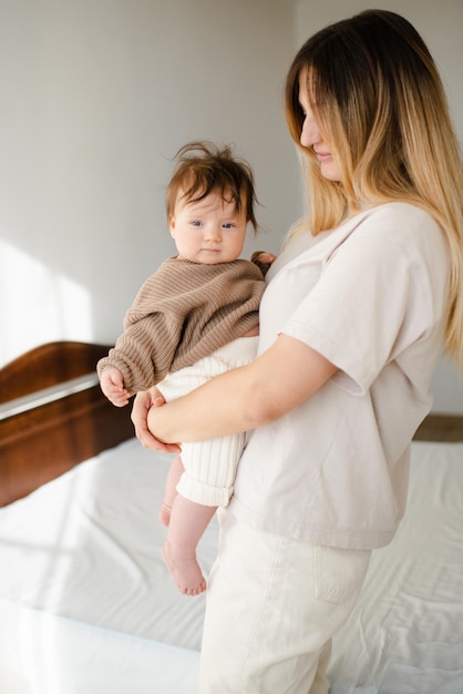Cute bebê menina com menos de 1 ano de idade em mãos de mãe usar suéter casual de tricô e calças no quarto Olhar para a câmera Maternidade