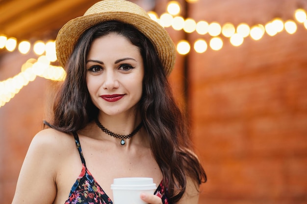 Cute brunette Frau mit dunklen Augen, roten Lippen und reiner gesunder Haut mit Strohhut und Kleid mit Kaffee zum Mitnehmen, der auf der Terrasse posiert Junge Frau mit Kaffeepause nach harter Arbeit im Büro