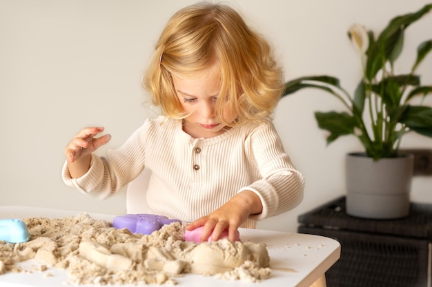 Cute happy caucasianblondecurlyhaired ToddlerBaby Girl spielt mit kinetischem Sand drinnenVorschulkind frühe Entwicklungmotorsensorische Fähigkeiten Konzept