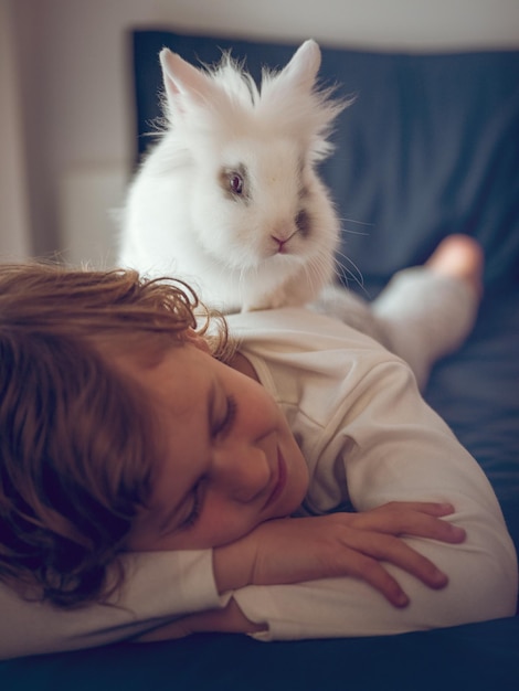 Cute Junge im Schlafanzug schläft friedlich auf einem bequemen Bett mit einem domestizierten weißen Kaninchen auf dem Rücken in einem hellen Schlafzimmer zu Hause