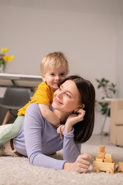 Cute little boy abrazando a su alegre madre mientras ambos están acostados en el suelo y jugando juguetes juntos en la sala de estar en cuarentena