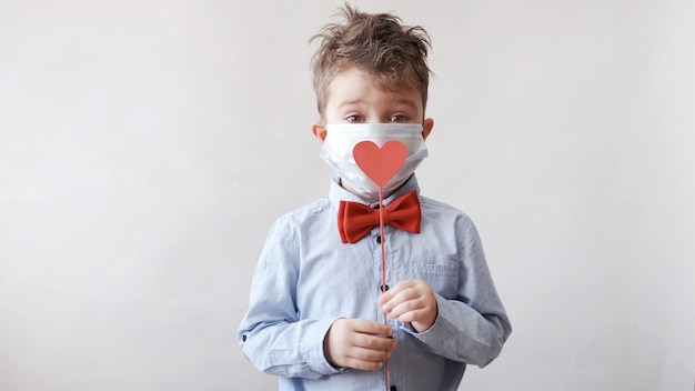 Cute little boy caucásico en pajarita con corazón de madera roja en palo en mascarilla protectora. día de San Valentín. COVID-19.