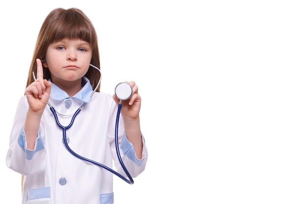 Cute little girl doctor vistiendo bata blanca sostiene el estetoscopio y muestra el gesto del dedo en la pared blanca aislada