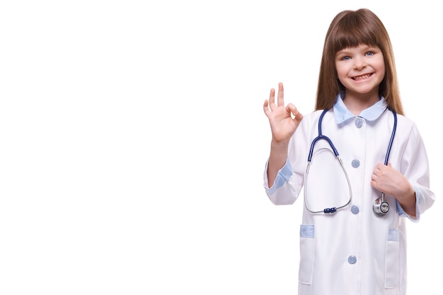 Cute Little Girl doctor vistiendo bata blanca sostiene un estetoscopio y muestra un gesto OK sobre fondo blanco aislado