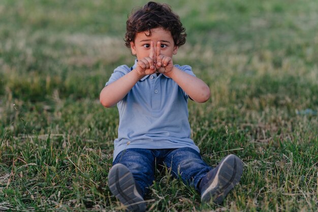 Cute lockiges kleines Kind schielt Augen verbunden seine Zeigefinger sitzt auf grünem Gras in Jeans und Polo gekleidet Lustige hispanische Kind spielt im Freien Kindheit und Freizeitaktivitäten für Kinder Gesunde kidsxAxA