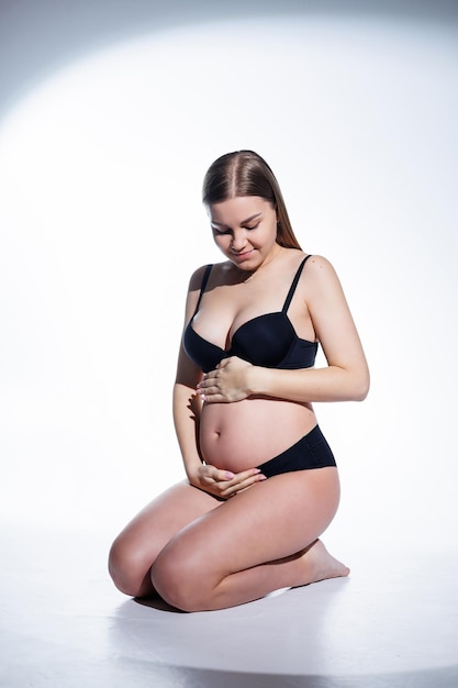 Cute schwangere Frau mit langen Haaren in schwarzer Unterwäsche sitzt auf einem weißen Hintergrund Damenunterwäsche für Schwangere