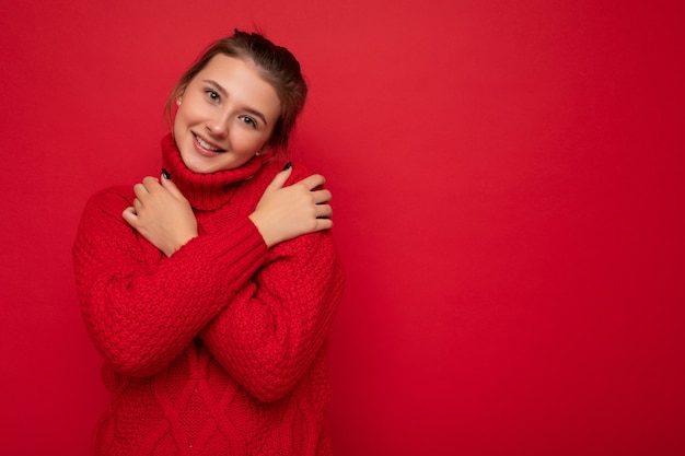 Cute sonriente joven vistiendo un cálido suéter rojo aislado en la pared roja