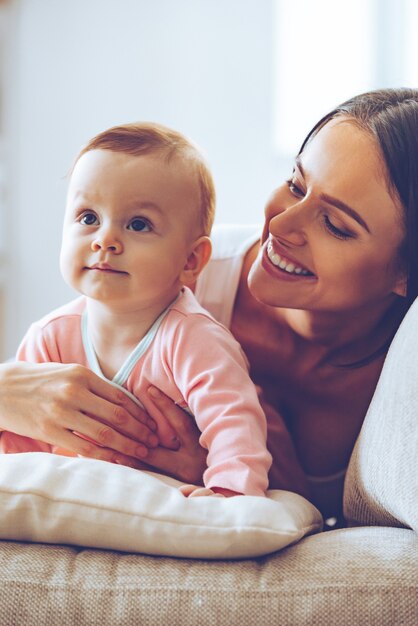 Cutie mit ihrer schönen Mama. Fröhliche junge Frau, die ihr Baby umarmt und sie ansieht, während sie zu Hause auf der Couch sitzt