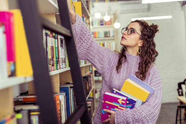 Da prateleira mais alta. jovem curiosa com um suéter de tricô roxo esticando a mão enquanto lê o livro gostei