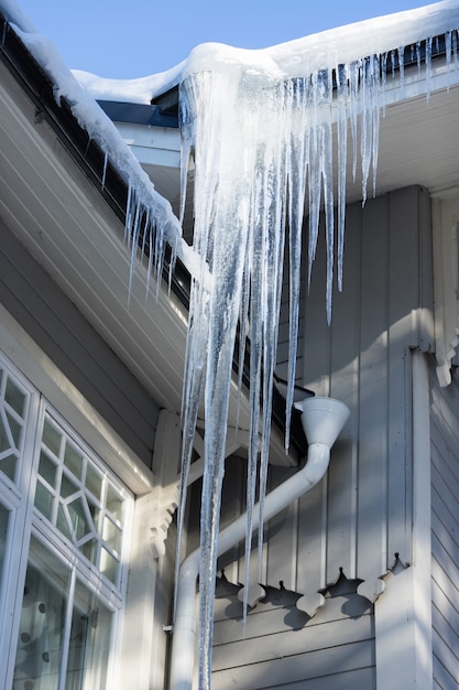 Dach des Holzgebäudes bedeckt mit scharfen Eiszapfen. Scharfer Eiszapfen, der an einem Abflussrohr hängt