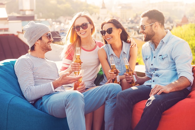 Dachparty mit Freunden. Vier junge fröhliche Leute, die sich unterhalten und Bier trinken, während sie an den Sitzsäcken auf dem Dach des Gebäudes sitzen