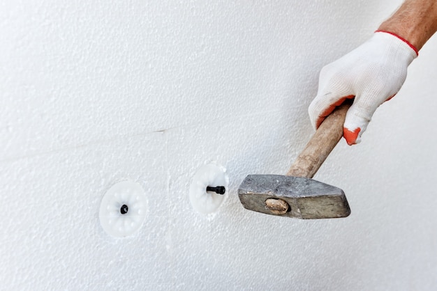 Foto dämmung des hauses mit polyschaum. ein arbeiter benutzt einen hammer für dübel, mit denen styroporplatten befestigt werden.