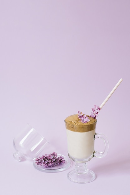 Dalgona Café em um copo transparente com um tubo de papel, um prato com flores lilás em um fundo lilás com um espaço de cópia