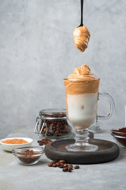 Dalgona Kaffee und Milch Koreanisches Getränk mit Eis und einem Löffel mit Schaum an einer grauen Wand. Ein erfrischendes, belebendes Getränk. Vertikale Ansicht.