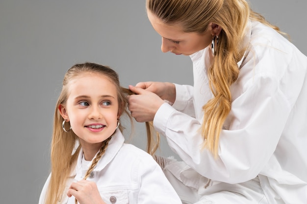 Dama adulta atenta con camisa blanca recogiendo el cabello de su hermana pequeña en trenzas finas mientras pasan tiempo juntos