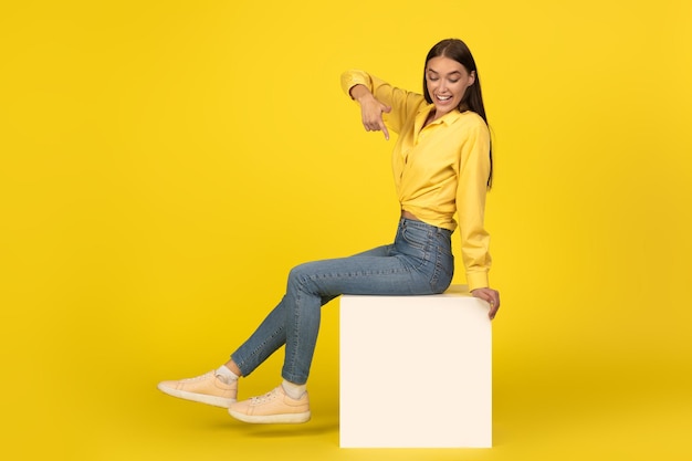 Una dama alegre sentada en un cubo señalando con el dedo el fondo amarillo