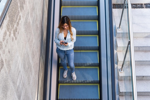 Foto dama alegre con teléfono inteligente caminando por la escalera mecánica mientras sonríe foto desde arriba