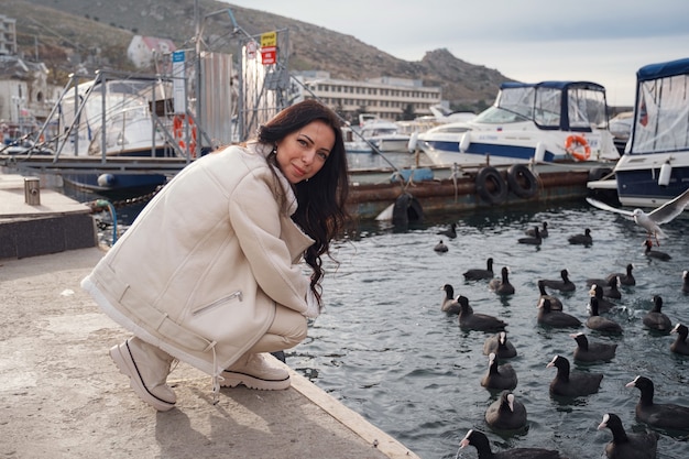 Una dama caucásica despreocupada con ropa beige disfrutando de la vista del mar en un día cálido y ventoso. Mujer alimentando patos y gaviotas en el paseo marítimo de la ciudad