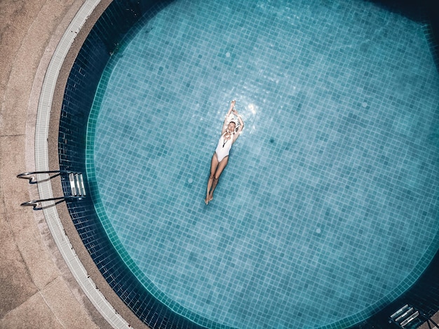 Una dama delgada y meditativa flota en la piscina azul redonda de un hotel tropical, con los brazos arriba, motas en el agua limpia. Concepto de moda, vista de drones