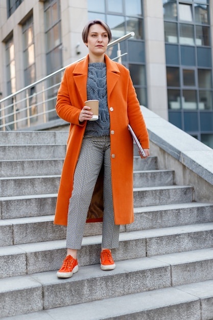 Dama elegante con abrigo naranja brillante en una escena de la ciudad, usando una computadora portátil y trabajando en ella durante un descanso. Tecnología al aire libre. tomando café, té, cacao