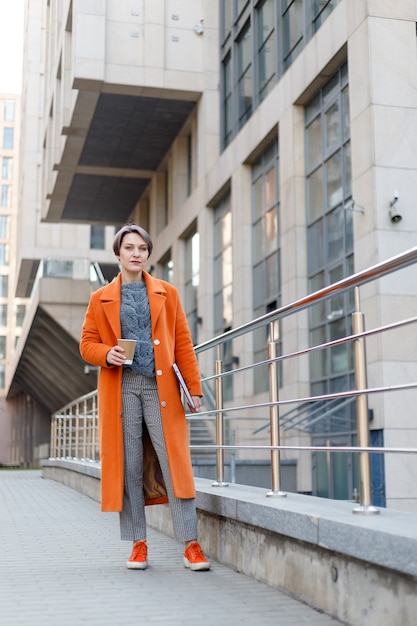 Dama elegante con abrigo naranja brillante en una escena de la ciudad, usando una computadora portátil y trabajando en ella durante un descanso. Tecnología al aire libre. tomando café, té, cacao