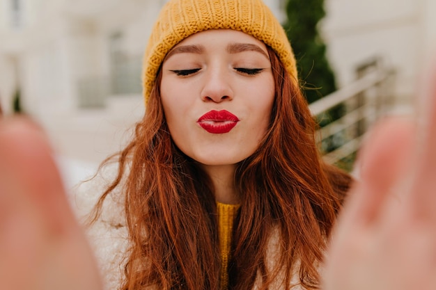 Dama elegante posando con expresión de cara de besos en el día de invierno Foto al aire libre de una chica de jengibre de ensueño con sombrero haciendo selfie