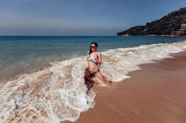 Una dama encantadora y sexy en bikini y gafas está arrodillada en la playa cerca del mar y las olas golpean su cuerpo. Concepto de aguas profundas.