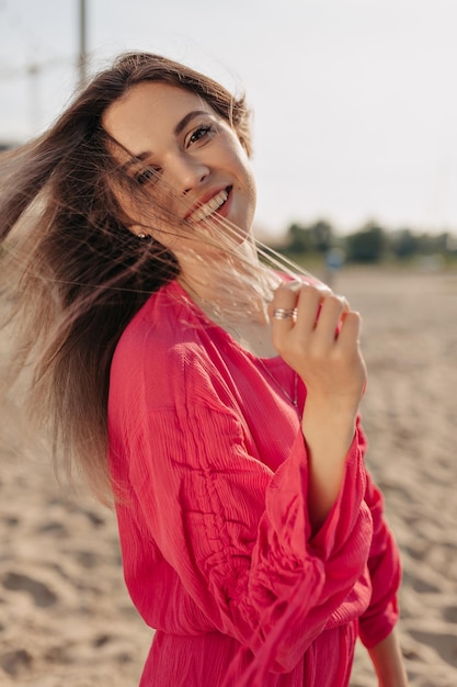 Una dama feliz y adorable con una amplia sonrisa se toca el pelo y se viste con una blusa rosa brillante en el fondo de la playa de arena