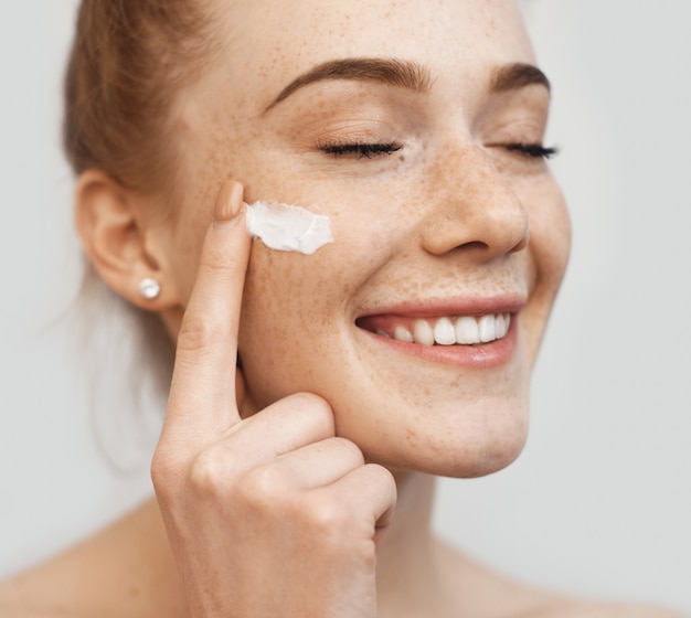 Dama de jengibre pecoso aplicando en la cara crema anti envejecimiento y sonrisa en una pared blanca de estudio