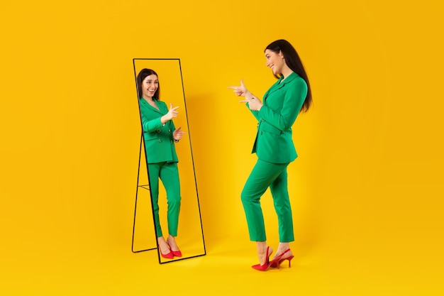 Dama de moda apuntando a su reflejo en el espejo posando después de una compra exitosa vestida de verde
