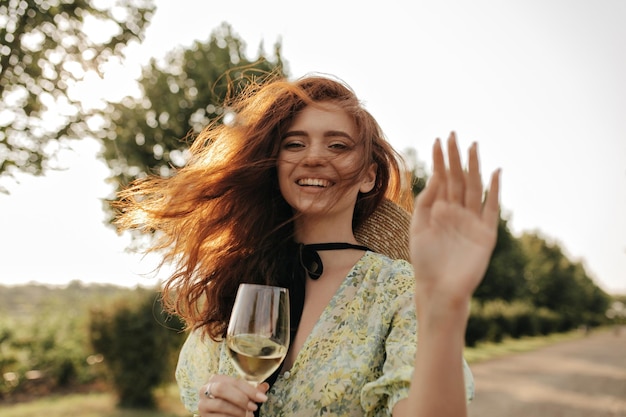 Dama moderna con cabello largo rojo en vestido estampado verde y amarillo riéndose mirando a la cámara y sosteniendo un vaso con vino al aire libre