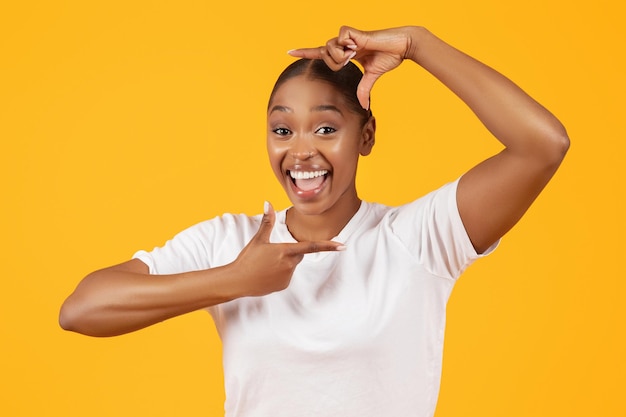 Dama negra con camiseta informal hace que los dedos enmarquen fondo amarillo