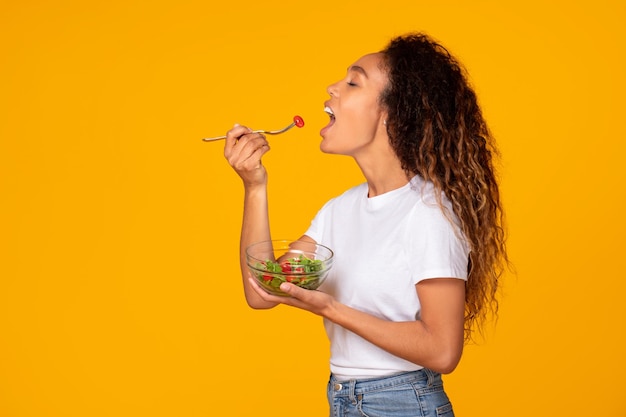 Dama negra delgada con un plato de ensalada comiendo fondo amarillo