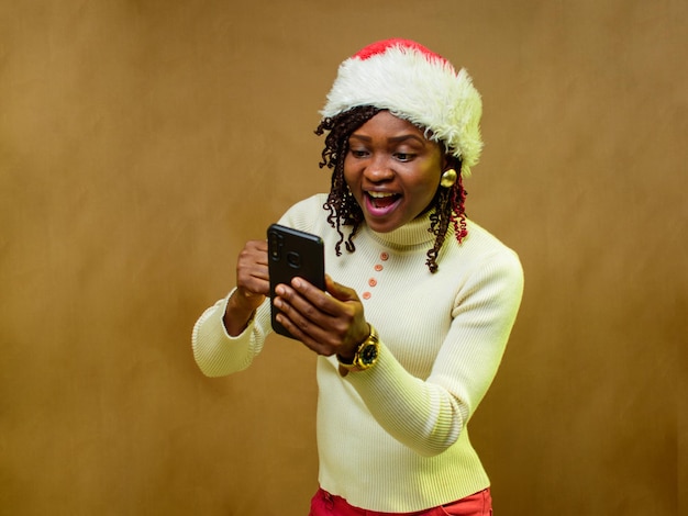 Una dama o mujer africana felizmente mirando el teléfono inteligente en su mano y también con un gorro de Navidad en la cabeza
