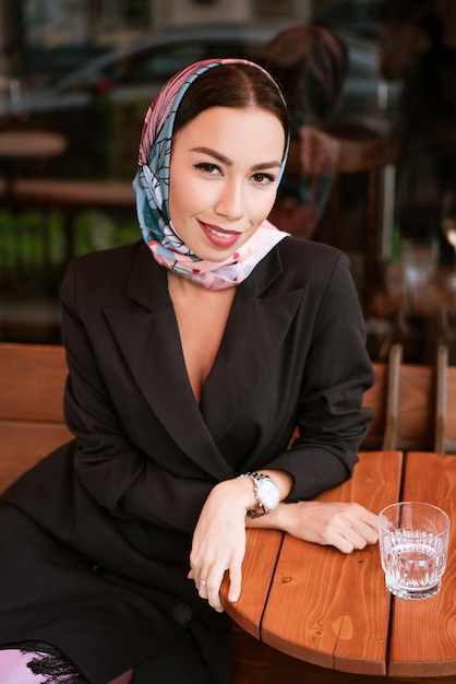 Una dama con un pañuelo en la cabeza y un vestido formal negro se sienta y sonríe en una mesa al aire libre. Concepto de reunión de negocios. De cerca.
