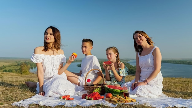 Una dama positiva con hijas e hijo se sientan juntos en la ribera verde y montañosa en un picnic festivo contra un cielo azul claro sin límites en un día soleado de verano