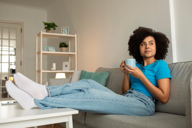 La dama rizada afroamericana milenaria pensativa disfruta relajándose y una taza de té en el sofá piensa mira