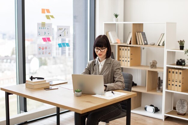 Dama de traje y gafas sentada cómodamente en la mesa con una computadora portátil