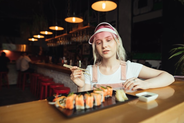 dama vestida de rosa sentada en una mesa en un restaurante asiático y comiendo apetitosos rollos de sushi