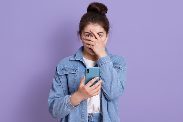 Dame, die Jeansjacke und weißes Hemd trägt, Mädchen mit brünettem Haar und Haarknoten, das Smartphone in der Hand hält und ihr Gesicht mit den Fingern bedeckt