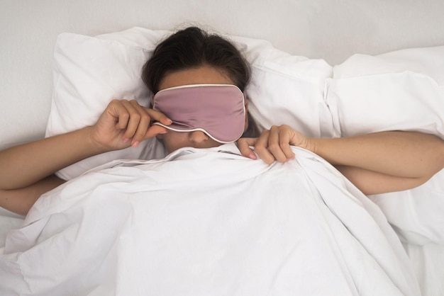 Dame mit dunklem Haar wacht auf und hebt farbige Schlafmaske auf Matratze mit heller Decke im Schlafzimmer