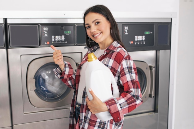 Dame mit Waschmittel gestikuliert mit dem Daumen nach oben in der Nähe von Waschmaschinen im Innenbereich