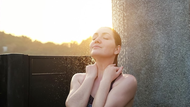 Dame steht unter der Außendusche des Hotelpools durch graue Betonwand gegen verschwommene Hügelsilhouetten unter hellem Sonnenaufgangslicht Nahaufnahme