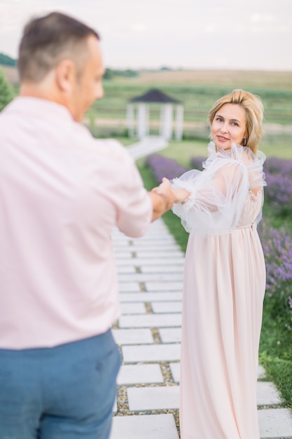 Dame tu mano, sígueme. Amor a través de los años juntos. Señora madura rubia bastante encantadora en vestido elegante, tomados de la mano y caminando con su hombre guapo al aire libre en campo lavanda con mirador