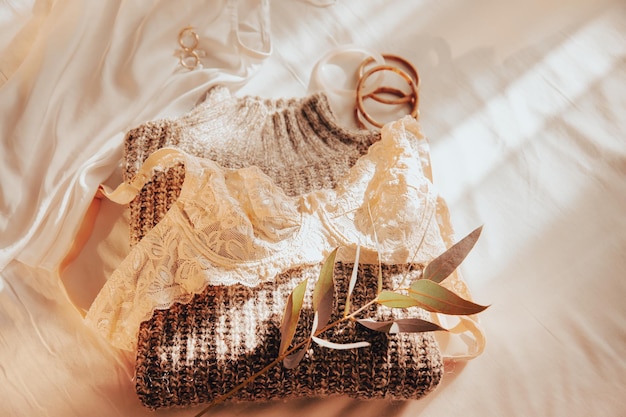 Damen Wintergarderobe Spitzen-BH Seidenoberteil Grobstrickpullover und Accessoires liegen in der Morgensonne auf einem zerknitterten Blatt