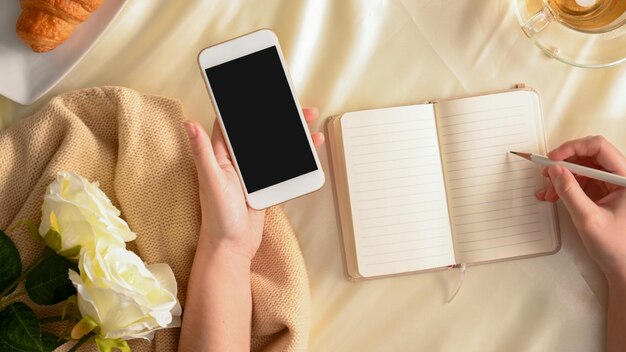 Damenschreibtisch Frau, die sich Notizen zu ihrem Buch macht und ein Smartphone über dem Arbeitstisch hält