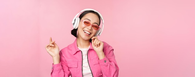 Dançando elegante garota asiática ouvindo música em fones de ouvido posando contra fundo rosa