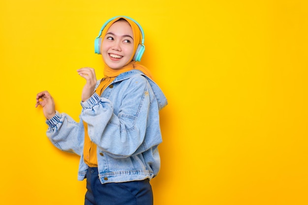 Dançando jovem asiática em jaqueta jeans usando fones de ouvido para ouvir música isolada sobre fundo amarelo