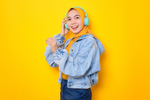 Dançando jovem asiática em jaqueta jeans usando fones de ouvido para ouvir música isolada sobre fundo amarelo